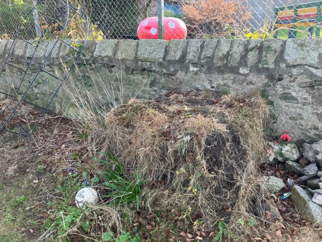 An overgrown compost heap and toppled archway with a weathered football nestled in weeds below a stone wall.