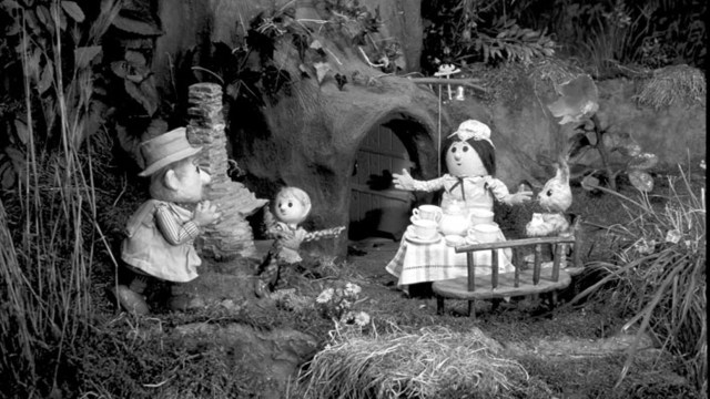 Black-and-white still from Pogle’s Wood showing Mr and Mrs Pogle, Pippin, and Tog outside their tree home with tea set.